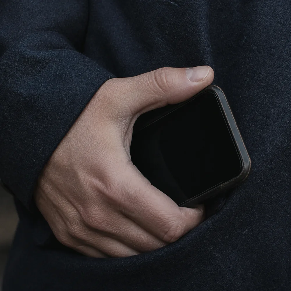 Close detail of a thumb resting on the edge of a phone with a dark screen, held against a wool coat.