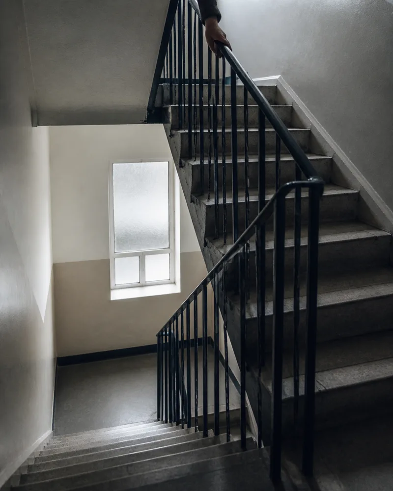 A communal stairwell in a British block of flats, worn stone steps and a painted handrail with one hand entering at the top of the frame.